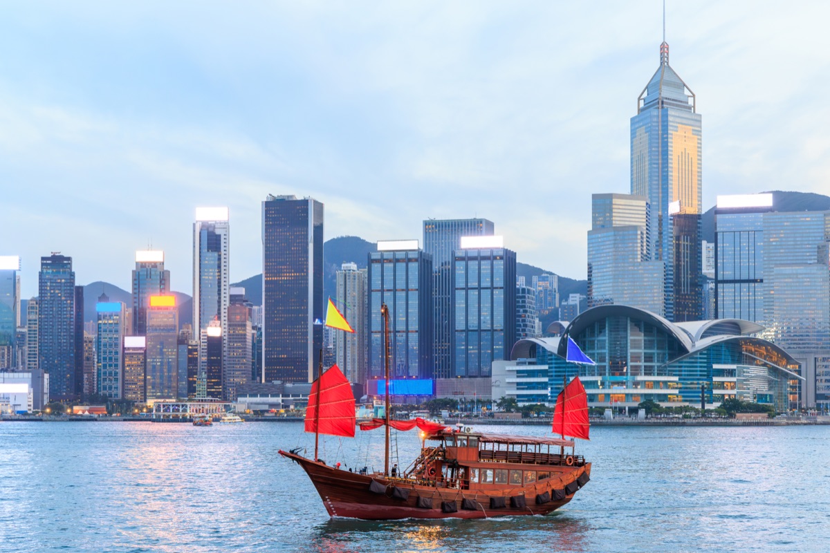 Traditional wooden junk boat with red sails against Hong Kong skyline during sunset, iconic urban scene