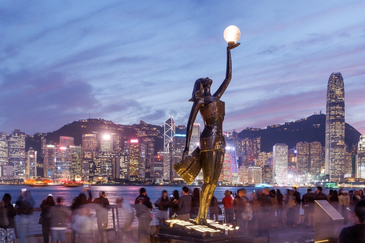 The Hong Kong Film Awards Statue with the city skyline in the background, bright night lights and crowds of tourists enjoying the colorful sunset