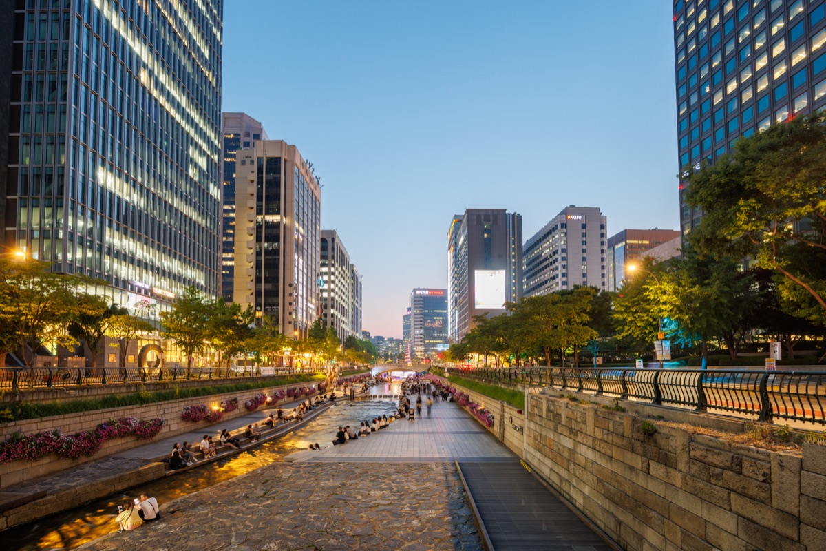 Visitors walk along Cheonggyecheon Stream surrounded by office buildings and modern city architecture in downtown Seoul.