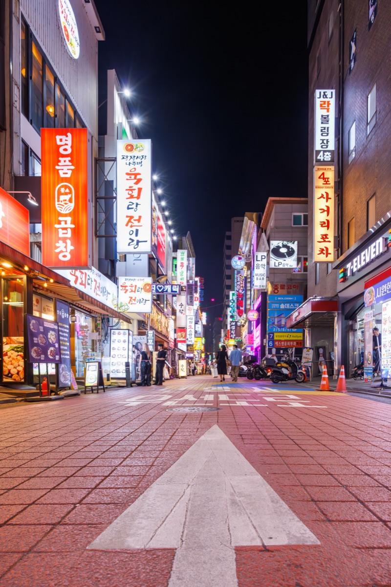 Illuminated storefronts and colorful signs along a pedestrian shopping street in the Myeongdong district of Seoul at night.