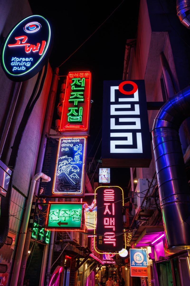 Bright neon signs illuminate a narrow entertainment alley in downtown Seoul at night