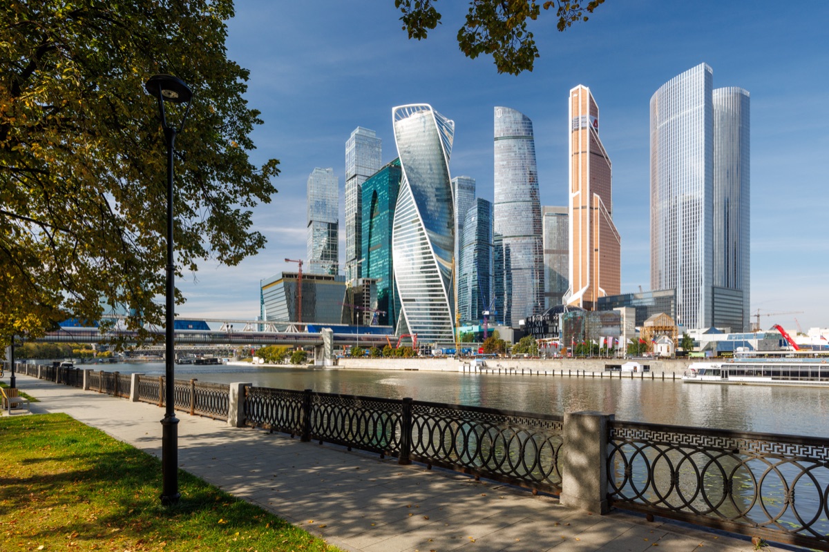 Moscow City towers rise across the river behind a decorative railing, with a street lamp and tree shadows on the walkway.