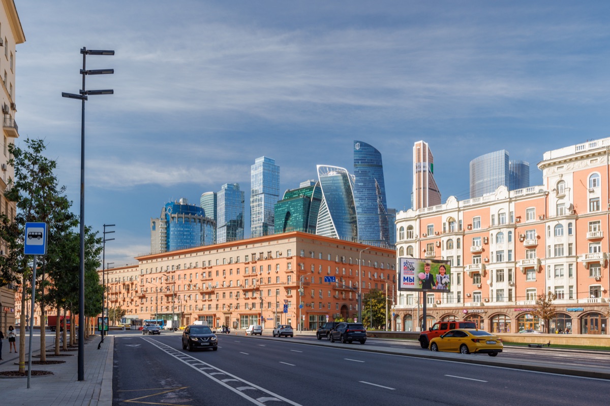 A yellow taxi passes older apartment blocks and a bus stop sign, with Moscow City skyscrapers rising in the background.