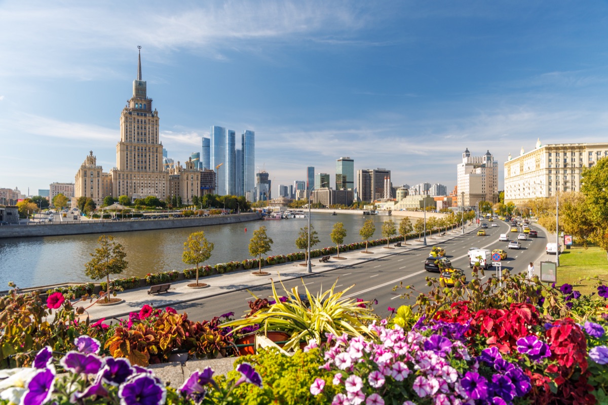 A broad view follows the Moskva River past Hotel Ukraina and distant skyscrapers, with road traffic and flower planters in front.