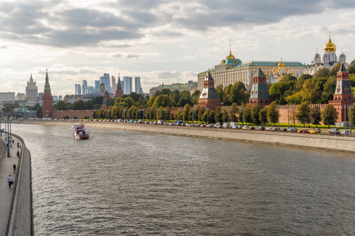 Cathedral domes and the Grand Kremlin Palace rise above the Kremlin walls, with boats on the river and distant skyscrapers beyond.