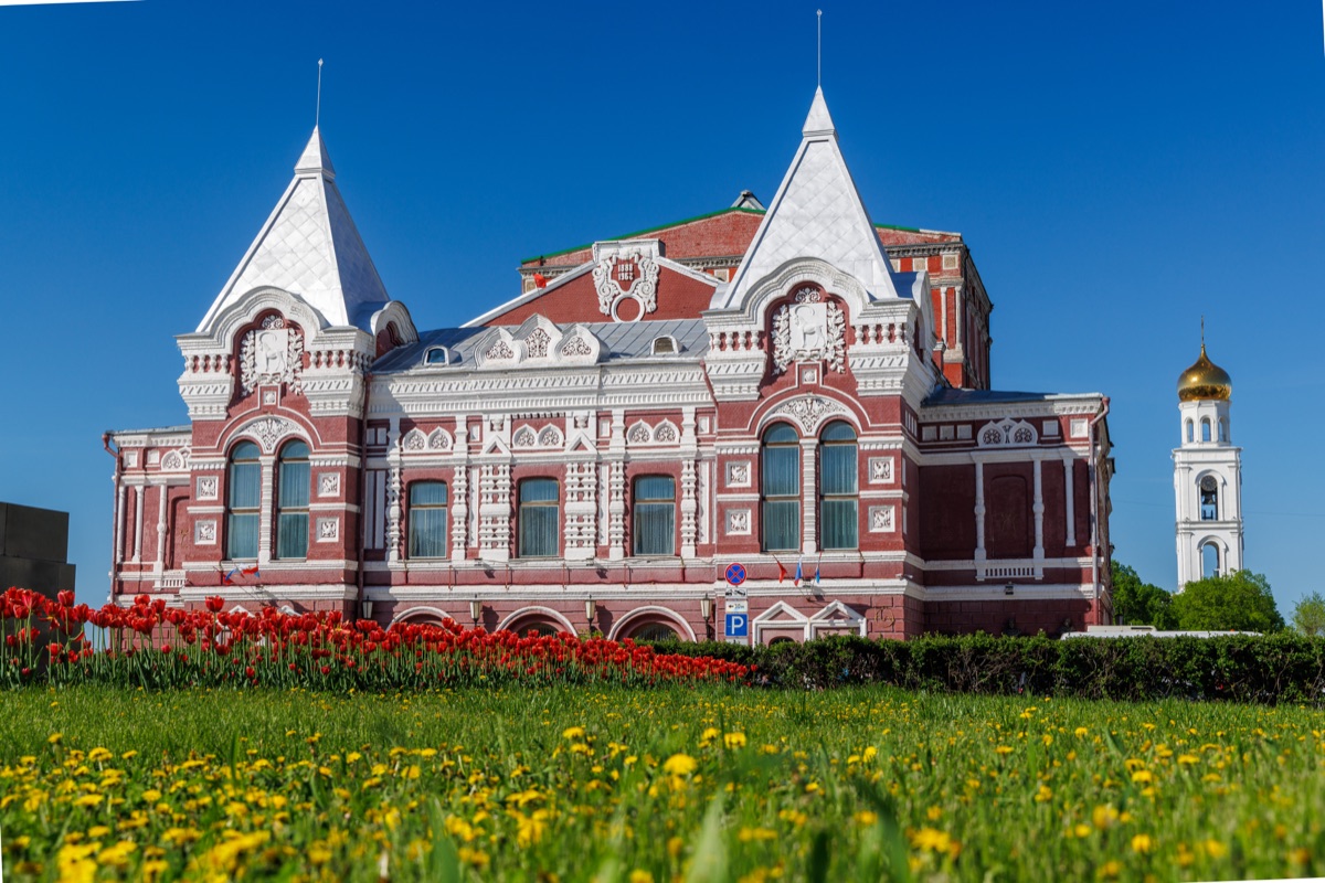 Samara Drama Theater building above green spring lawn with yellow dandelions under blue sky