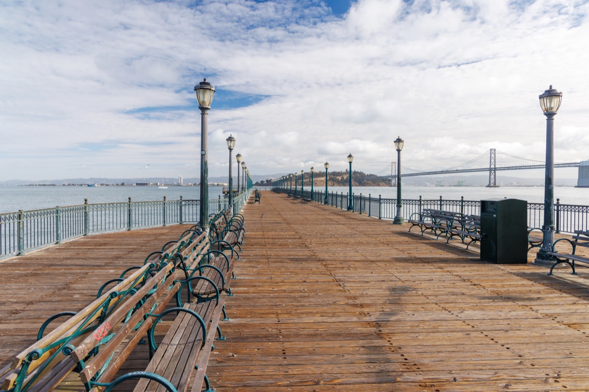 Scenic view of a classic wooden pier stretching into the San Francisco Bay, framed by calm waters and iconic city landmarks in the distance, under a bright sky
