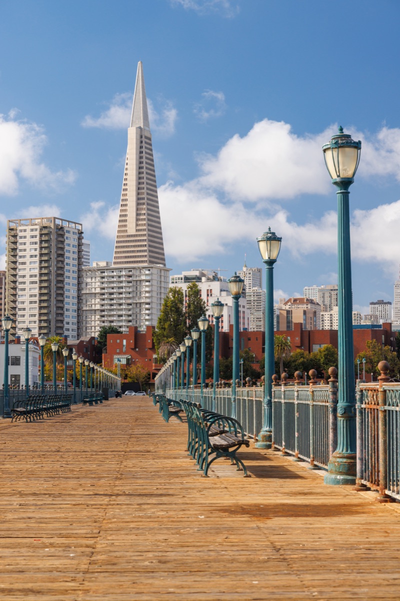 Scenic view of a classic wooden pier stretching into the San Francisco Bay, framed by calm waters and iconic city landmarks in the distance, under a bright sky